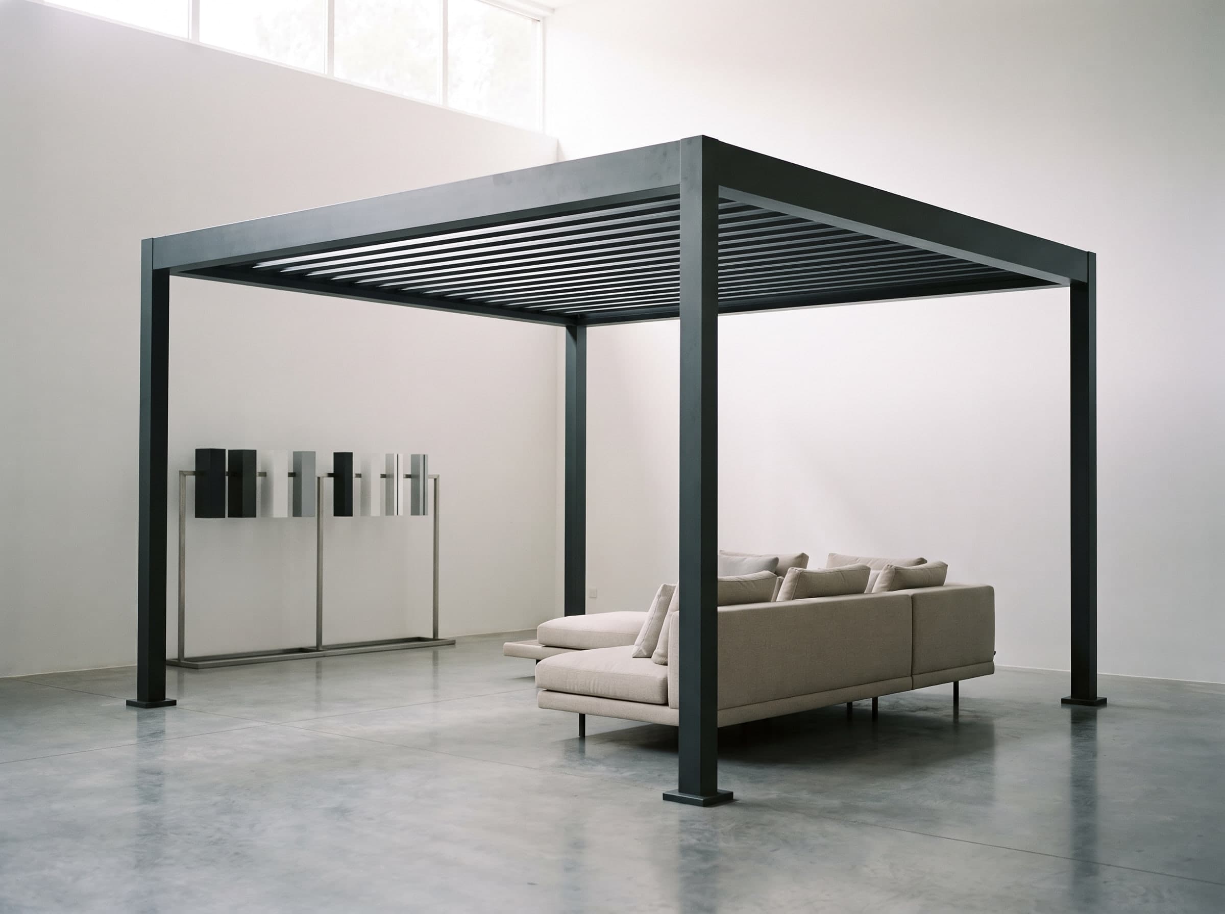 Free-standing anthracite louvred pergola on display in a bright showroom with a sofa and colour samples